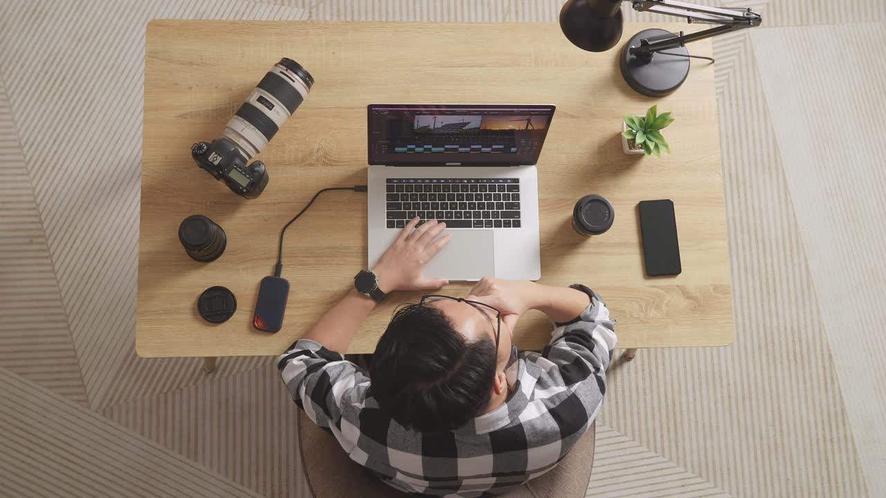 vista superior de un editor masculino asiático pensando en las imágenes mientras está sentado en el espacio de trabajo usando una computadora portátil junto a la cámara editando el video en casa
