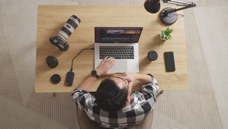 top view of asian male editor thinking about the footage while sitting in the workspace using a laptop next to the camera editing the video at home