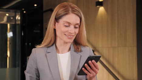 happy satisfied adult business woman standing in office hall looking into mobile phone smartphone screen chatting online in net smiling answering message in social media using gadget device.