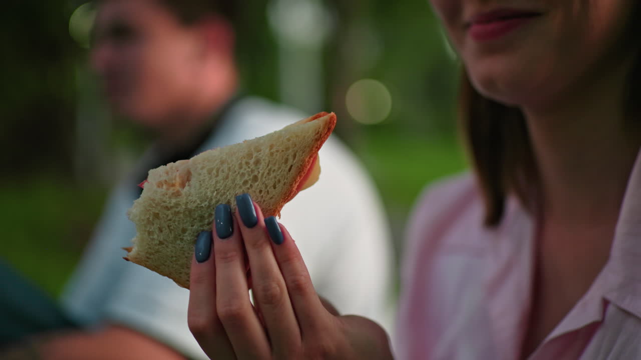 close-up de una mujer con uñas pulidas observando un sándwich a la parrilla en su mano, con un fondo borroso que muestra a alguien masticando sándwich en un entorno al aire libre con iluminación suave