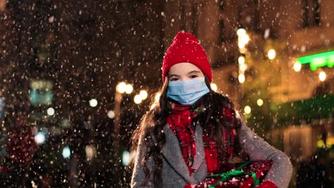 portrait of happy caucasian little girl in scarf holding a present and smiling at camera while it¬¥s snowing on the street in christmas