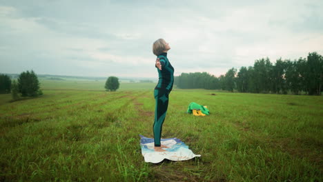 side view of middle aged woman in green and black suit and leggings doing yoga on a mat in grassy field, slowly moving her hands up and down in a peaceful practice, in vast grassy field