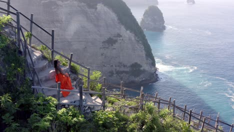 kelingking beach in nusa penida island indonesia with woman in orange dress sitting and looking at paluang cliff, aerial flyover reveal shot