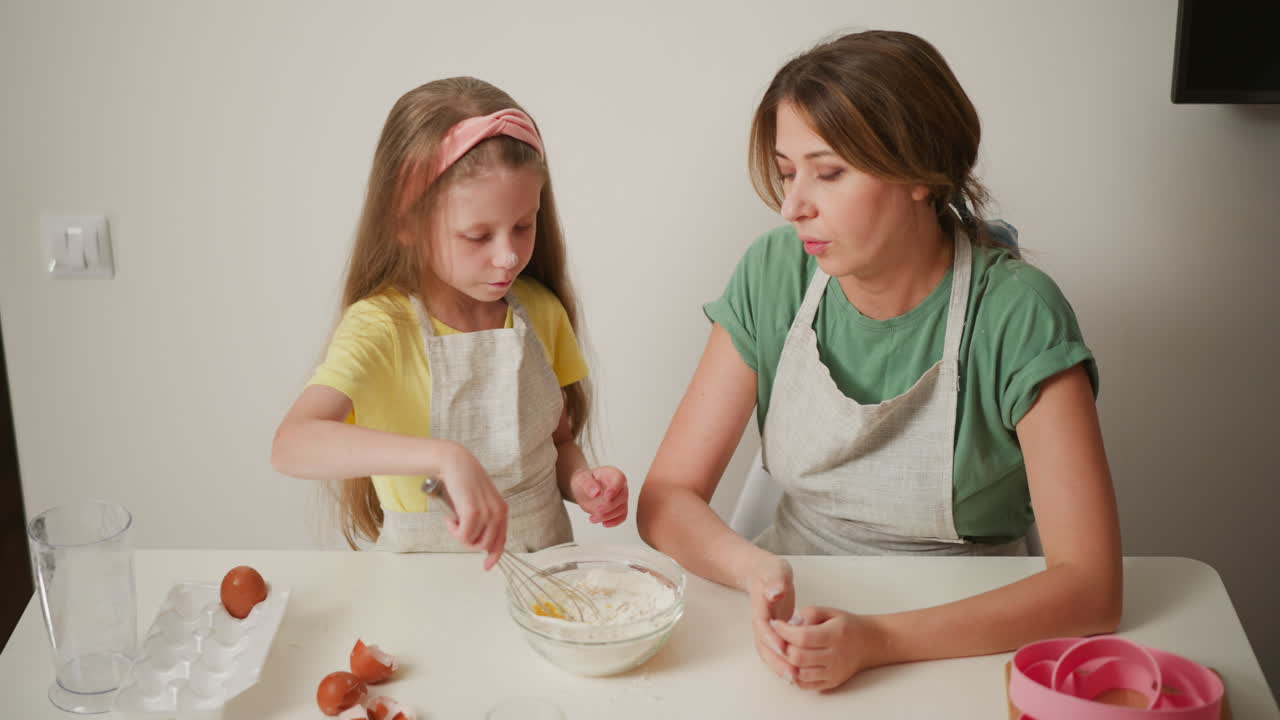 young girl wearing apron whisking flour and egg mixture in glass bowl while woman in green shirt and apron watches beside her, flour stains on table and cracked eggshells scattered near bowl