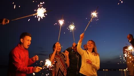 um grupo de jovens amigos a fazer uma festa na praia. amigos a dançar e a celebrar com faíscas no pôr-do-sol do crepúsculo.