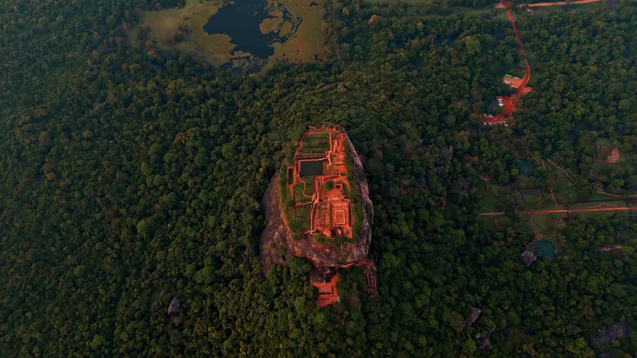 Top-down vertical drone shot over Sigiriya Rock Fortress at sunrise, revealing the ancient symmetrical layout of the ruins surrounded by dense jungle and glowing morning mist.