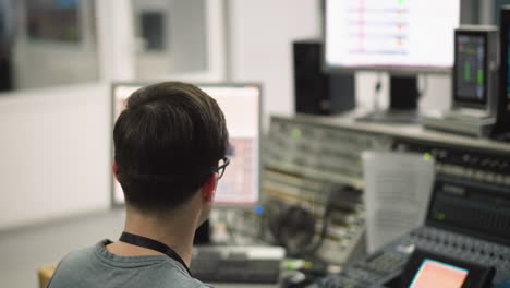 broadcast operator seated at sound desk adjusting equipment, focused on work. monitors display live video feed, radio and television production in studio environment
