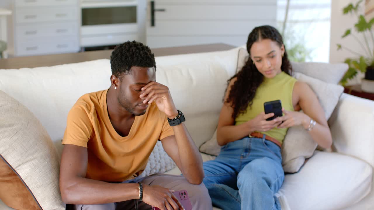 Casal afro-americano sentado no sofá, o homem esfregando a testa enquanto a mulher mexe no celular.