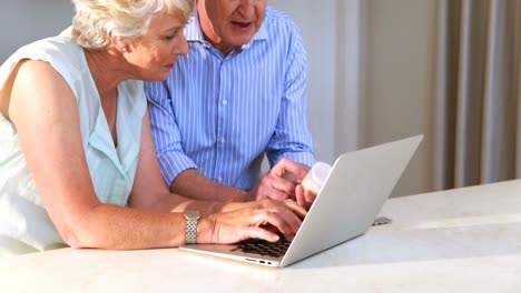 una pareja de ancianos comprobando la medicina mientras usan una computadora portátil en la cocina