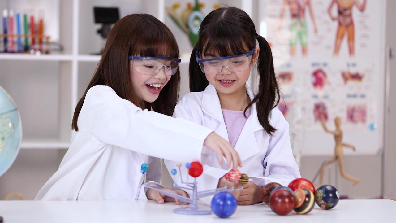 Two girls wearing safety goggles examine a solar system model in a bright science classroom.