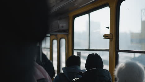 back view inside moving train shows people in winter coats and beanies seated while others stand, soft daylight through window, urban commute mood, worn metal interior, and frost outside glass