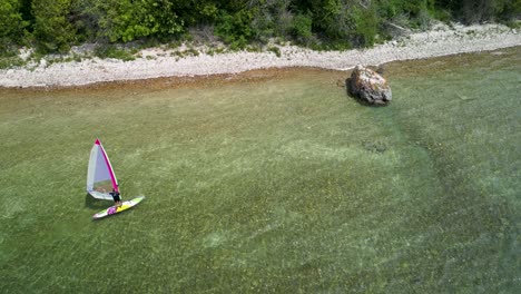 Aerial-view-of-windsurfer-arriving-at-large-rock-on-shoreline,-Lake-Huron,-Michigan