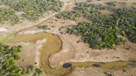 Flying-over-a-dry-river,-across-trees-and-fields