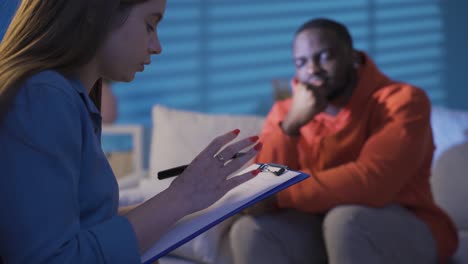 female psychologist listening to black man with psychological problems and making notes on paper.