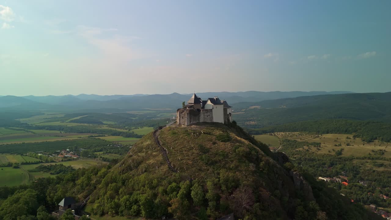 Aerial view of Füzér Castle perched on a hill, surrounded by lush greenery, rolling hills, and a scenic countryside landscape in Hungary's Borsod-Abaúj-Zemplén region