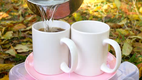 a person makes coffee outdoors. he pours water from the jug into the camping cup and stirs with a spoon. against the backdrop of the autumn landscape.