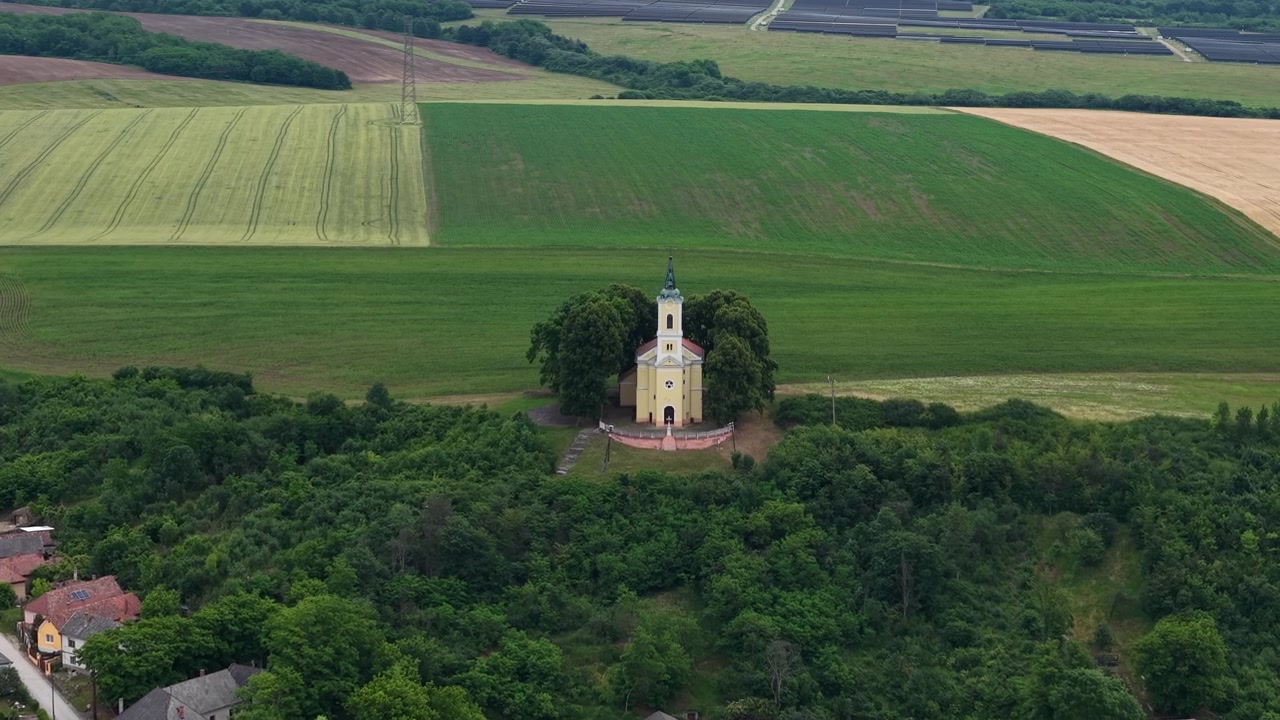 En historisk ungersk kyrka står på toppen av en grön kulle, med utsikt över en charmig lantlig by, vidsträckta fält och en avlägsen solpark