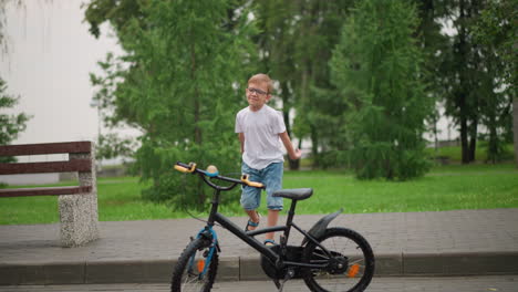 a young boy playfully walks toward a parked bicycle, raising his hands in the air before grabbing the handlebars, dressed in a white top and denim shorts