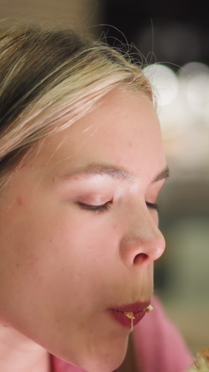 mujer de vestido rosa toma un bocado de hamburguesa mientras sostiene papas fritas, comiendo con una expresión satisfecha, el fondo presenta una vista borrosa de una persona de rojo caminando y una vista de luz borrosa
