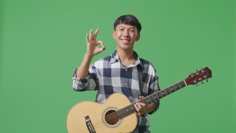 asian teenager with guitar showing okay gesture and smiling on green screen background