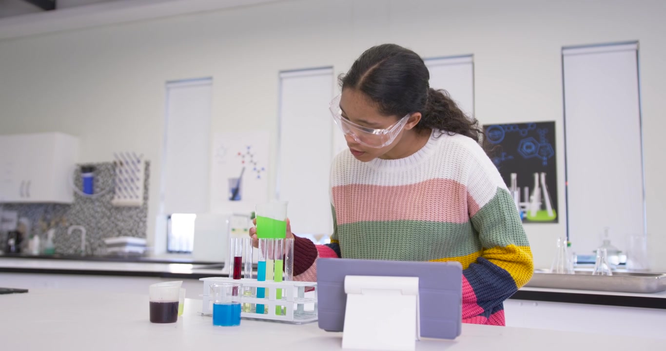 Schoolgirl putting on goggles checking tablet at lab placing green cup in rack, examining liquids