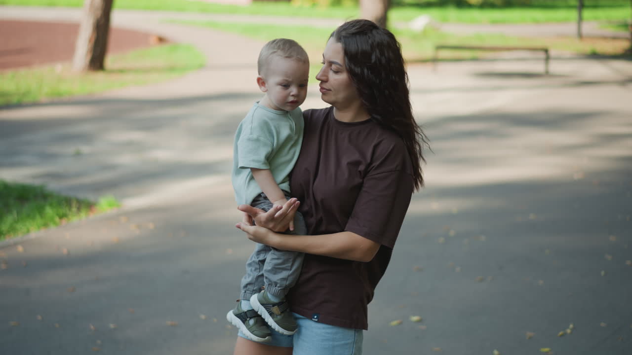 Madre latina che tiene in braccio un bambino nel parco, primo piano intimo che mostra una dolce rassicurazione, sorriso soffice e abbraccio protettivo, momento spontaneo all'aperto che evidenzia una genitorialità affettuosa e il legame quotidiano.