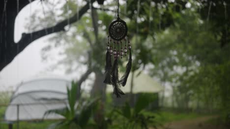 Un-Atrapasueños-Cuelga-Delicadamente,-Capturando-La-Esencia-De-La-Tranquilidad,-Mientras-La-Lluvia-Cae-Con-Gracia-En-Un-Paisaje-Verde-Denso-Y-Exuberante