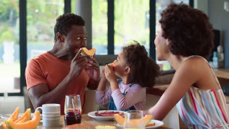 Familienaufnahme-Mit-Eltern-Und-Tochter-Zu-Hause,-Die-Vor-Dem-Frühstück-Lustige-Grimassen-Mit-Melone-Schneiden