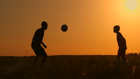 Vater-Und-Sohn-Spielen-Fußball-Im-Park-Bei-Sonnenuntergang,-Silhouetten-Vor-Dem-Hintergrund-Einer-Zeitlupenaufnahme-Einer-Hellen-Sonne.