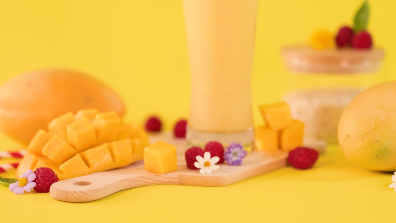Slow-motion spoonful of cream drops beside mango smoothie, fresh fruit, and yogurt on yellow