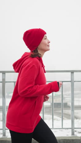 side view of woman jogging in red beanie and jacket along snowy park pathway near iron railing overlooking bridge with passing cars in foggy winter atmosphere showing visible breath