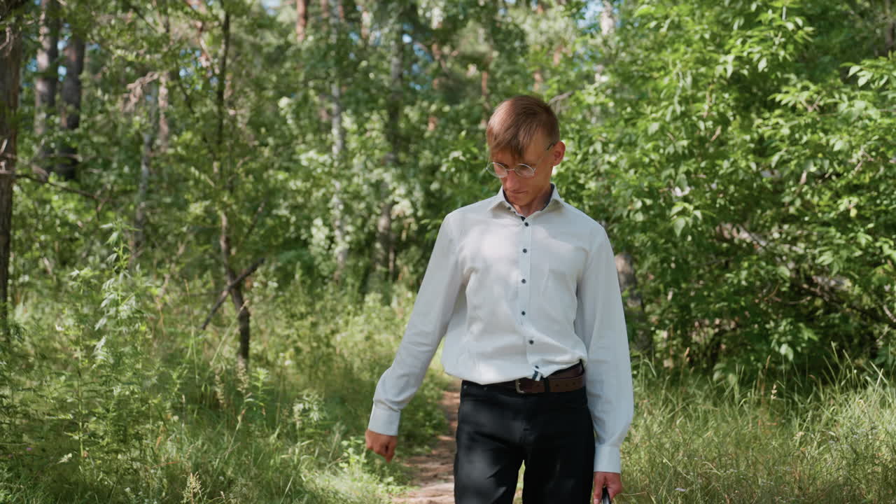 Tall handsome man in white top and black trouser carrying black backpack walking slowly through forest path, carefully observing plants along way surrounded by lush greenery