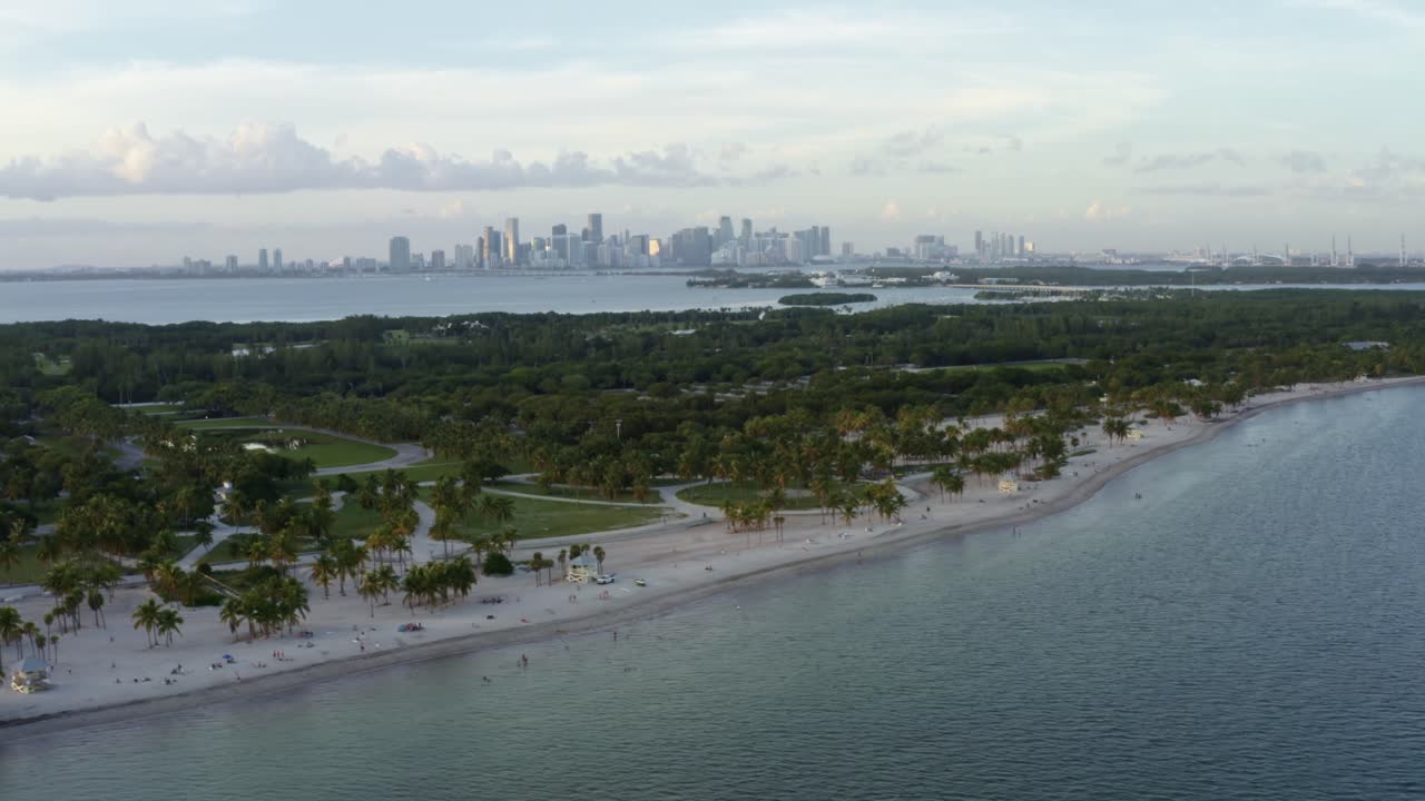 왼쪽을 기울이는 트럭 항공 드론 촬영 열대 해변, 나무로 둘러싸인 크랜던 파크, 키 비스케인, 플로리다, 마이애미의 스카이 라인, 멀리, 은 여름 저녁