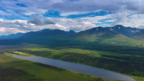 Approaching a long pond among the green meadows. Fluffy white clouds hang over the mountains at backdrop. Alaska aerial