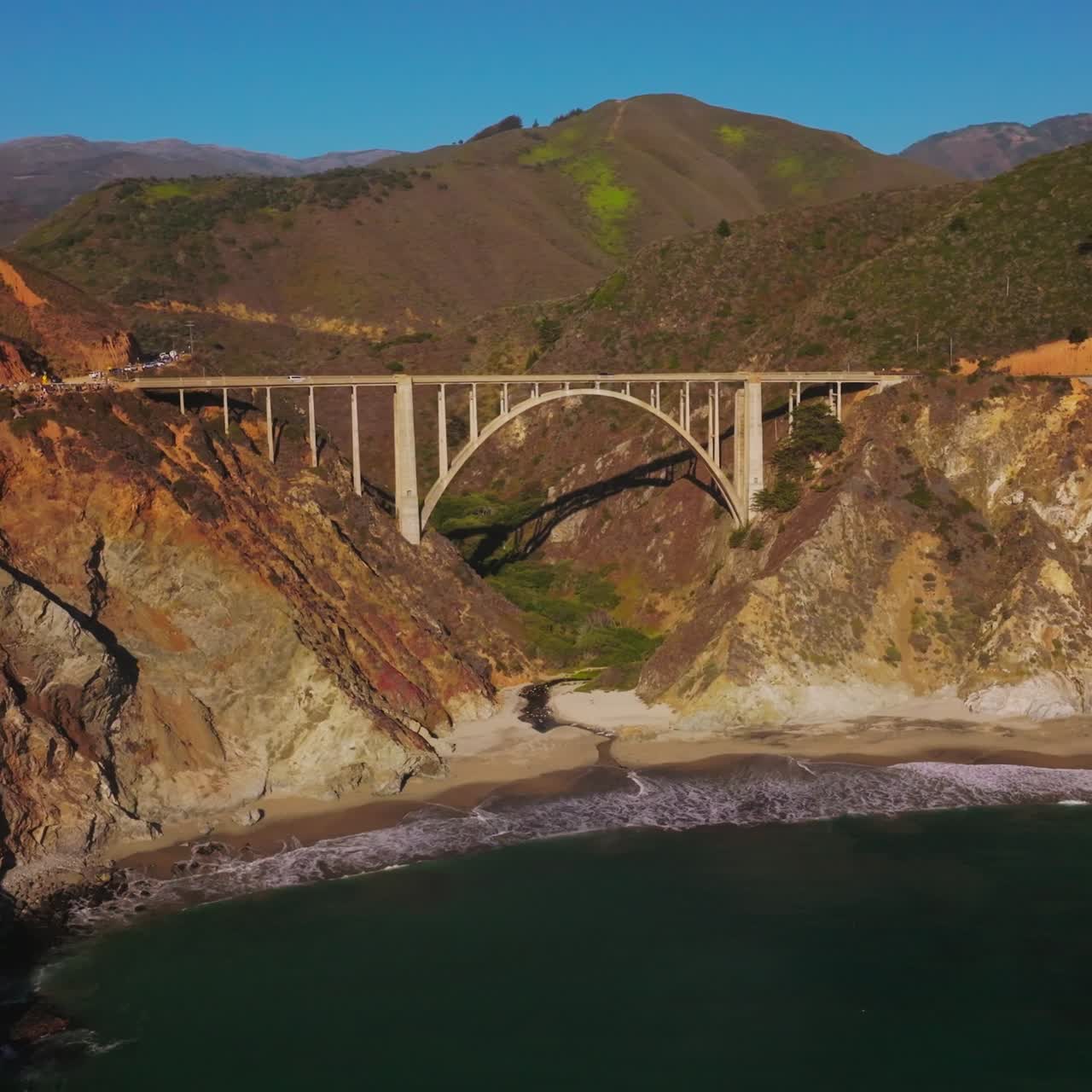 Beautiful sight of rocky shore with thin line of a sandy beach at Pacific coast. Arched bridge locating between two mountains with automobile road on it