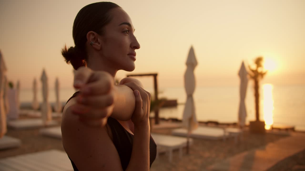 A brunette girl with tied hair in a black top does stretching of the upper body and warms up her shoulders before doing sports on a sunny beach during the sunrise