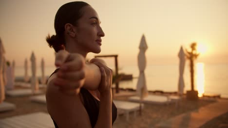 a brunette girl with tied hair in a black top does stretching of the upper body and warms up her shoulders before doing sports on a sunny beach during the sunrise