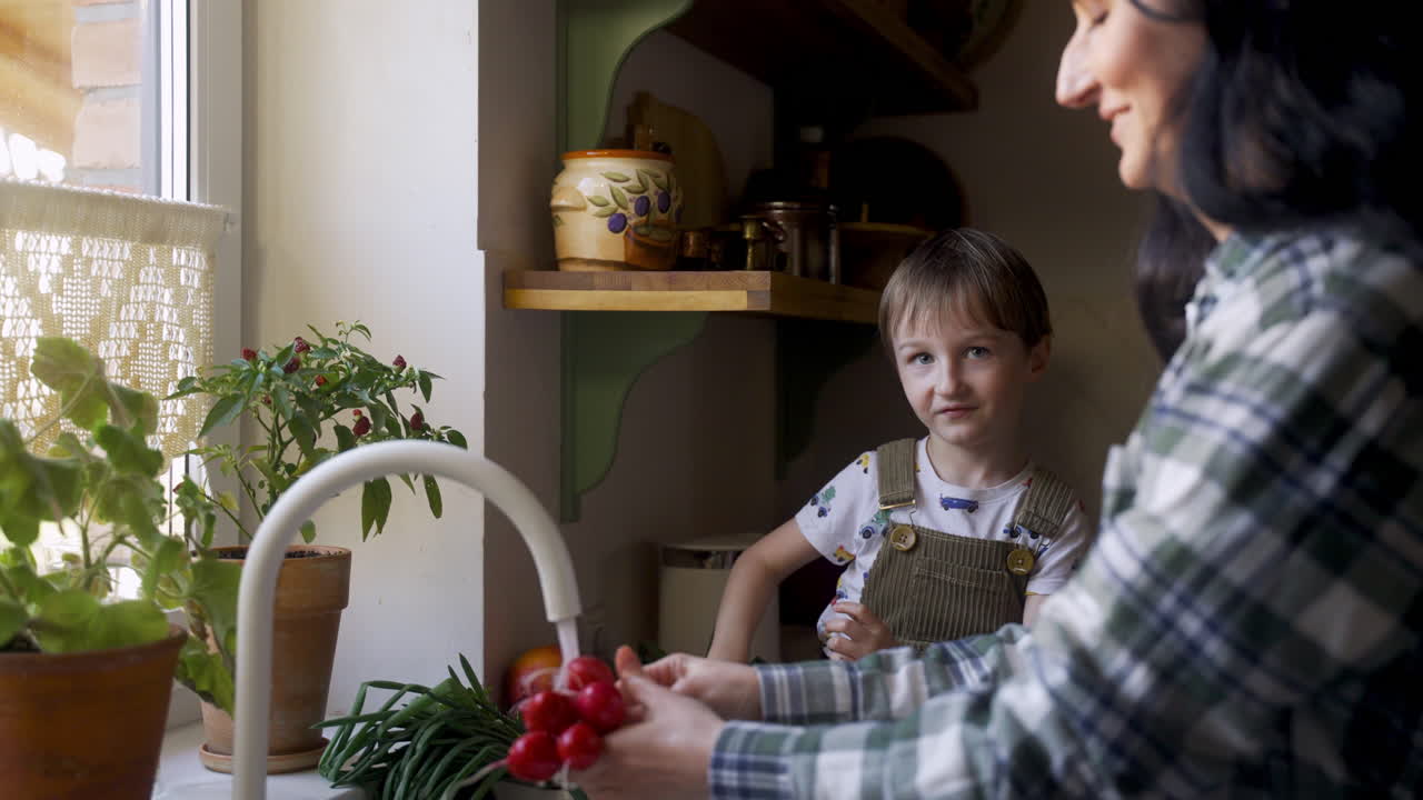 vista lateral de una mujer caucásica lavando verduras y frutas en el fregadero. su hijo la ayuda