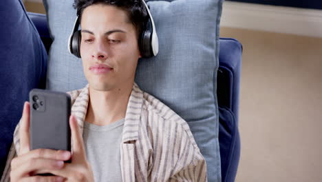 Happy-biracial-man-in-headphones-lying-on-couch-using-smartphone,-copy-space,-slow-motion