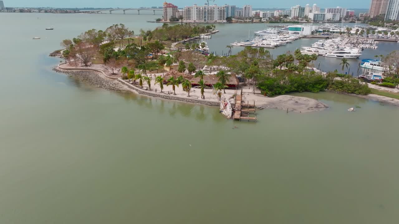 Aerial reverse dolly shot over grounded sailboat and mangled dock, revealing Sarasota Bay, Marina Jack, and downtown Sarasota. Grounded boats, debris, and birds visible post-Hurricane Milton.