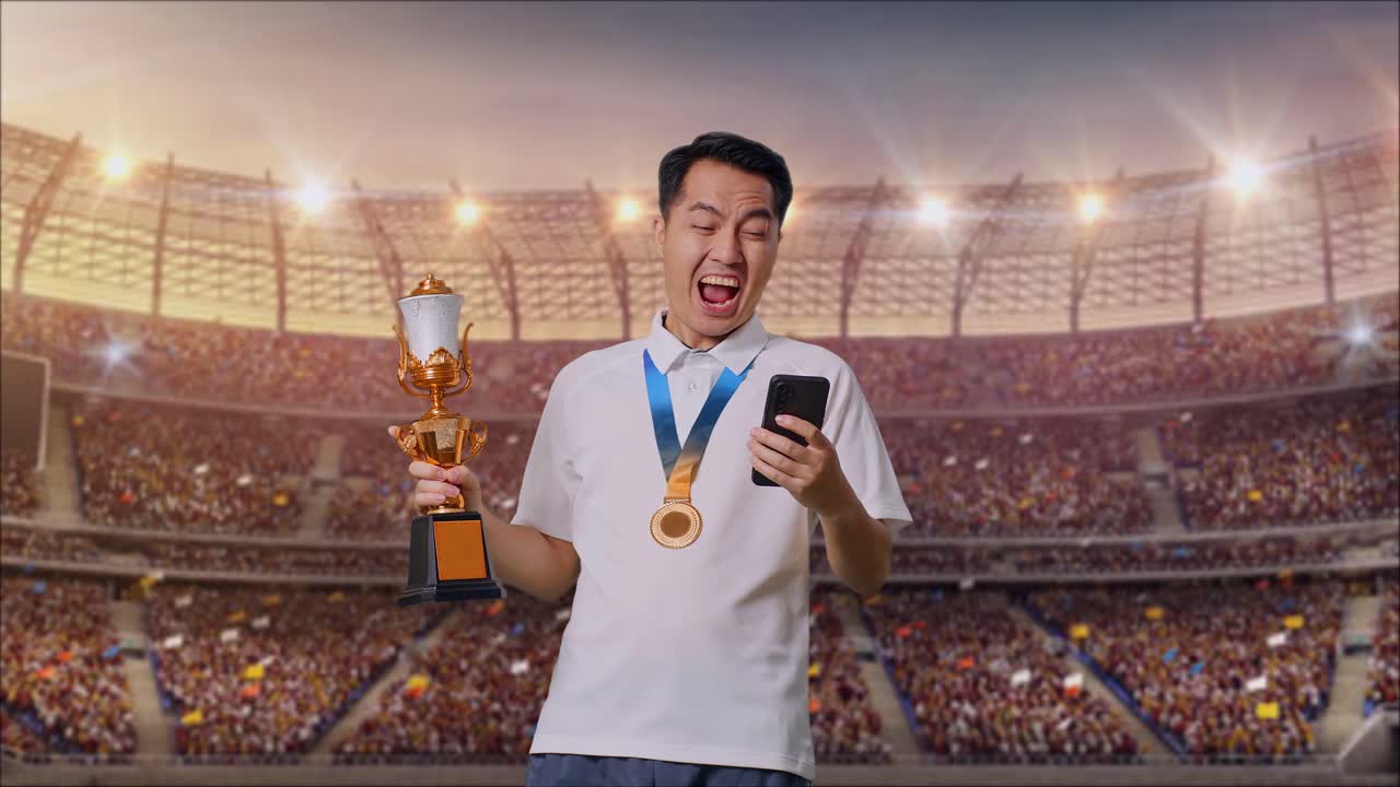 금메달과 트로피를 가진 아시아인 남자가 스마트폰을 보고 경기장에서 골을 축하하고 소리쳤다.
