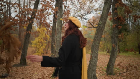 lady in black coat, yellow beret, and yellow muffler touches dry foliage, looking around with excitement in vibrant autumn forest covered with fallen leaves