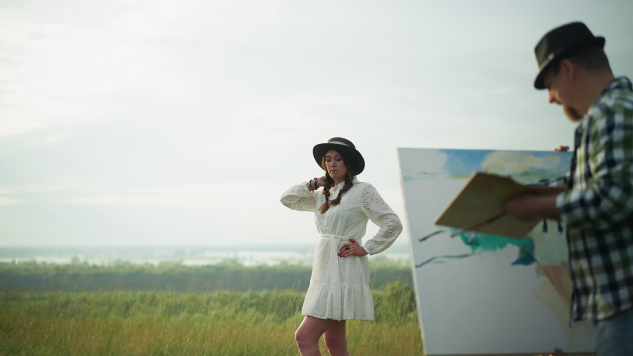 A woman in a white dress and hat poses elegantly in a grassy field while an artist in a checkered shirt and jeans, holding a palette, paints her portrait