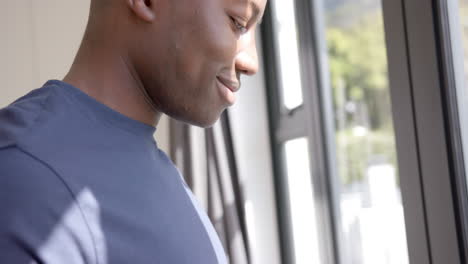 african american man using smartphone and looking through window at home, slow motion