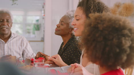 multi generation family hold hands around table at home saying grace before eating christmas meal