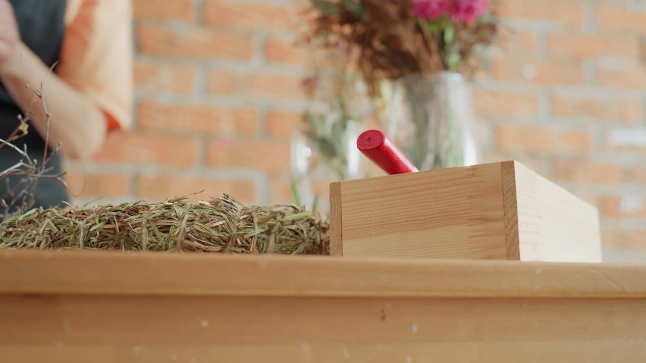 Wooden box with red handle placed on wooden table beside bundle of dry hay and blurred background of flowers in glass vase against brick wall in rustic workshop interior with natural decorative materials