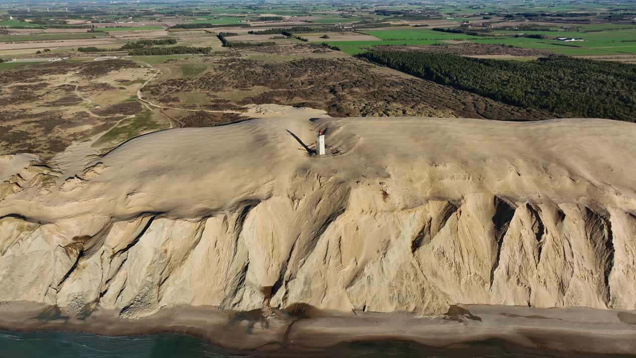 Experience a stunning drone panoramic view of Denmark’s coast, where rolling sand dunes meet a timeless stone lighthouse, blending natural beauty with historic charm in one captivating scene.