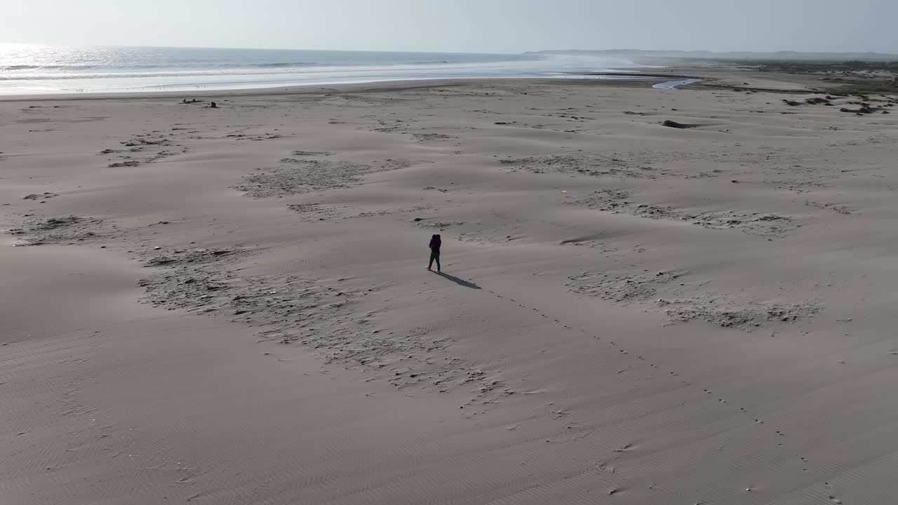 페루의 칼레타 비달 해변에서 모래사장을 걷는 사람의 공중 패닝 샷은 화창한 날 배경에 잔잔한 파도와 도시가 내려다보이는 바다를 내려다보고 있습니다.