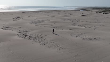 Toma-Panorámica-Aérea-De-Una-Persona-Caminando-En-La-Arena-De-La-Playa-De-Caleta-Vidal-En-Perú-Con-Vistas-Al-Mar-Con-Olas-Tranquilas-Y-La-Ciudad-Al-Fondo-En-Un-Día-Soleado