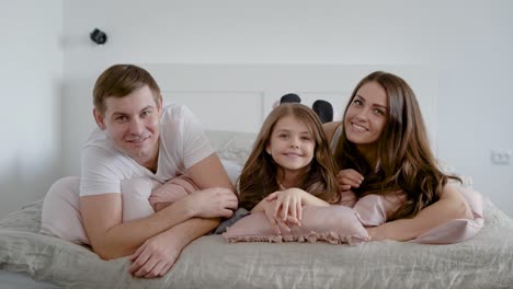 happy little girl is lying on pillow on bed between her father and mother, mom is kissing daughter in cheek in home